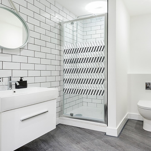 Sticker format carré adhésif motif chevrons horizontaux blanc et noirs collé à la porte de la douche d'une salle de bain éclairée et moderne